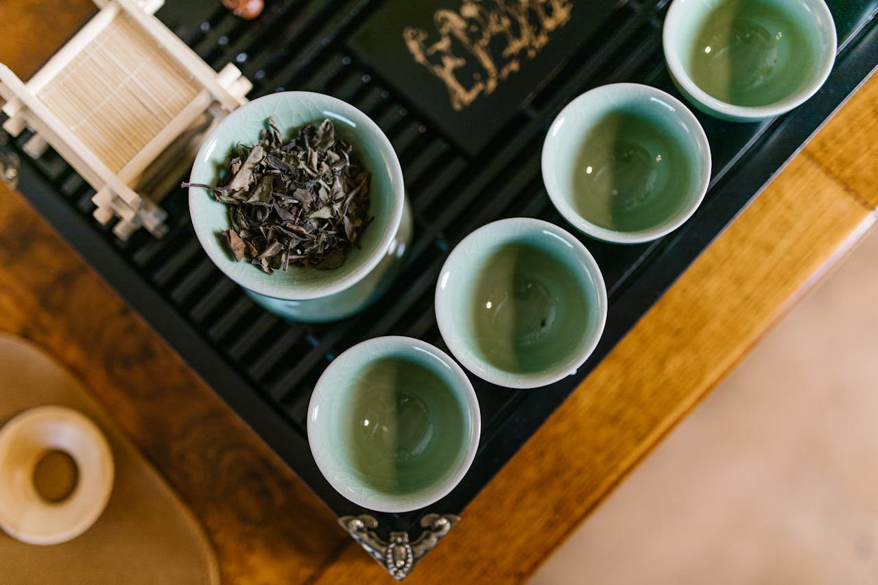 Top view of a traditional tea set with green teacups and dried tea leaves on a bamboo rack, captured in a warm setting.